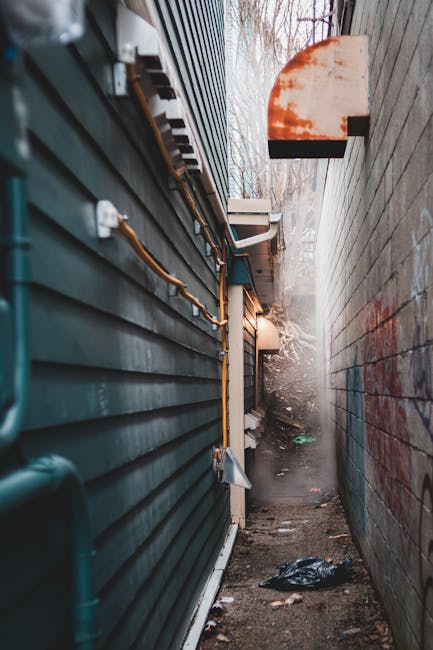 A large, weathered red metal dumpster with visible rust and chipped paint is positioned against a dark green tiled wall and a light grey concrete wall on a paved urban street. Several clear plastic ba