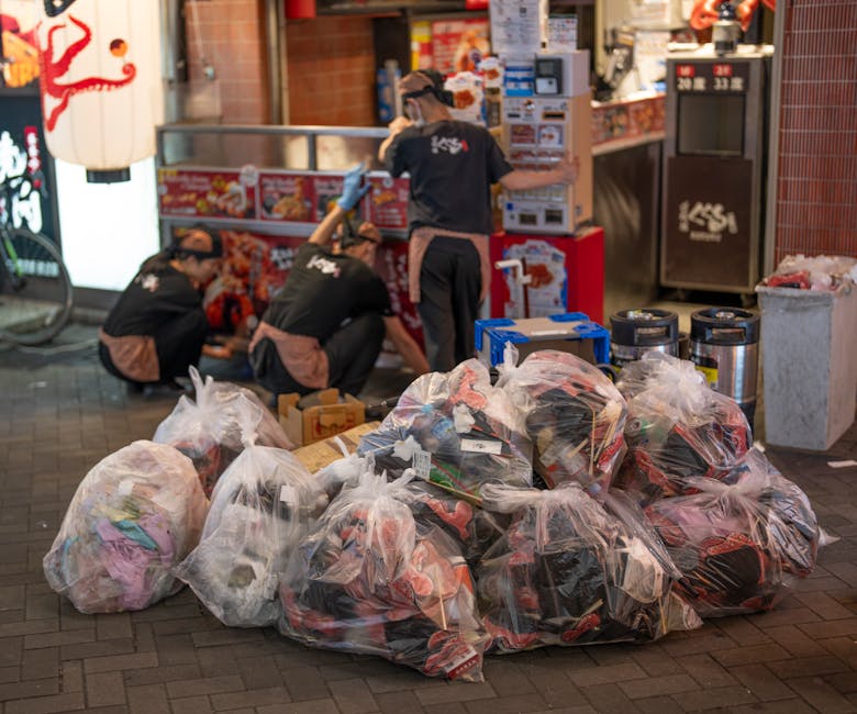 A collection of large transparent plastic bags filled with assorted discarded clothing and textiles is arranged on the pavement outside a commercial establishment at night. The plastic bags are bulgin