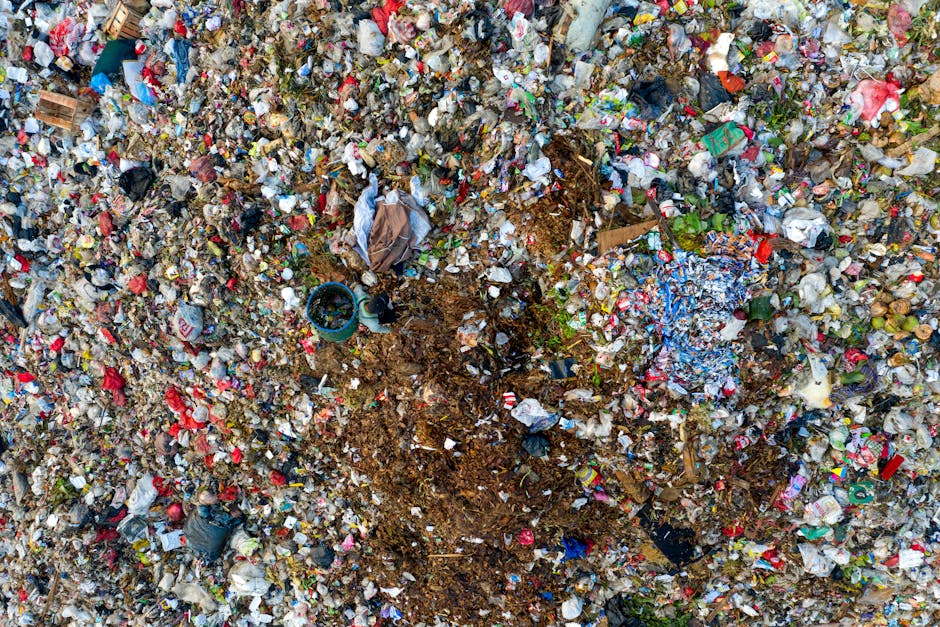 A large open landfill site covered with a substantial accumulation of various types of waste, including discarded tires, plastic bottles, cardboard boxes, and fabric materials. The tires are scattered